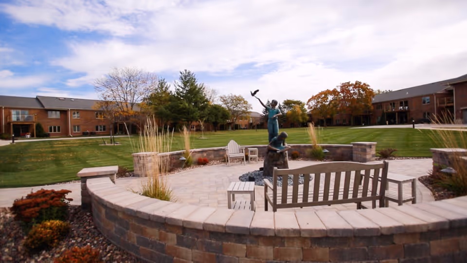 Outdoor courtyard area at Tudor Oaks Senior Living Community featuring a circular stone seating wall, wooden benches, chairs, and a bronze statue of two children playing with a bird. The courtyard is surrounded by well-maintained green lawns and two-story brick buildings under a partly cloudy sky.