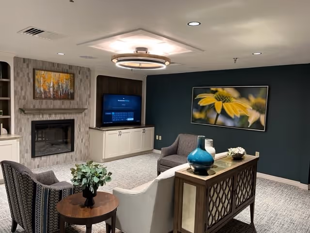 A cozy living room area with a fireplace on the left wall, a flat-screen TV on a cabinet, two armchairs, a small round wooden table with a plant, and a console table with decorative vases. The walls are painted dark green and light gray, and there is a large framed photograph of a yellow flower on the wall.