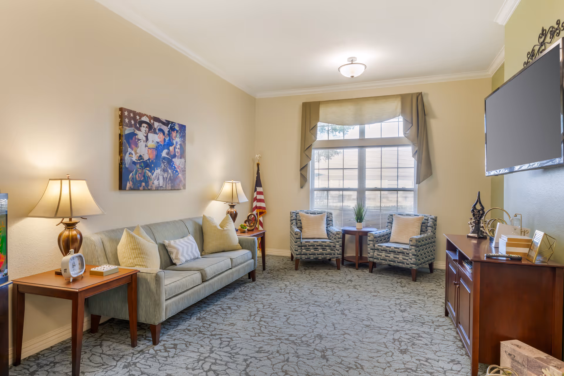 A cozy living room with a light green sofa adorned with several pillows, two patterned armchairs with pillows, a wooden side table with a lamp and clock, and a wooden TV stand with decorative items. A large window with blinds and a valance lets in natural light. A patriotic-themed painting and an American flag are displayed on the walls.