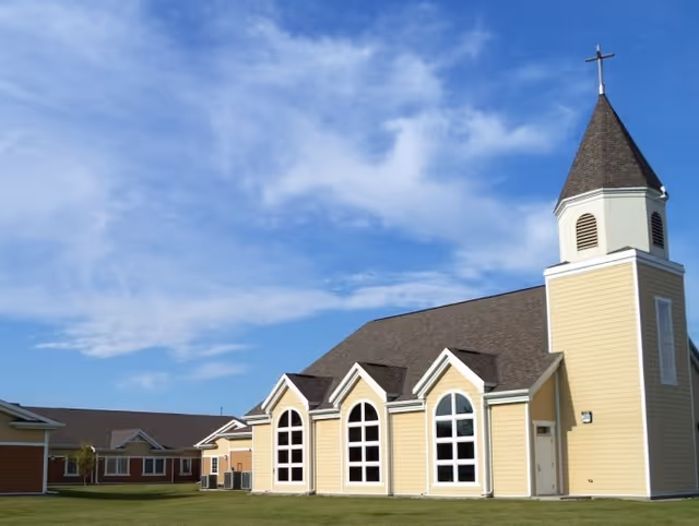 Exterior view of a yellow building with a steep roof and a tall steeple topped with a cross, under a partly cloudy blue sky. The building has large arched windows and is surrounded by a grassy area.