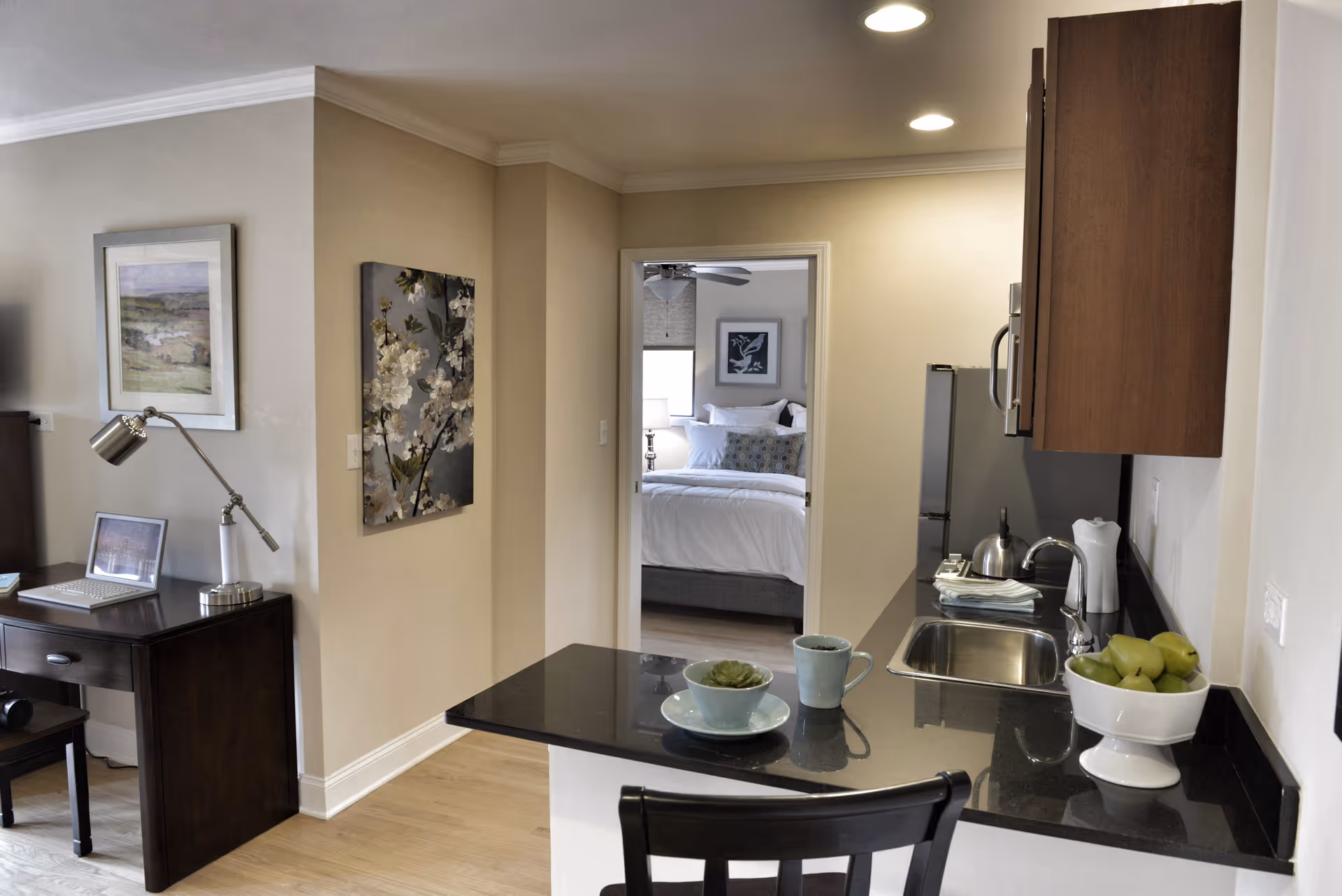 Interior view of a senior living facility showing a small kitchen area with a black countertop, a sink, a bowl of green apples, and a cup with a saucer. To the left, there is a dark wooden desk with a lamp and a laptop. In the background, a bedroom with a neatly made bed and framed artwork on the wall is visible through an open doorway.