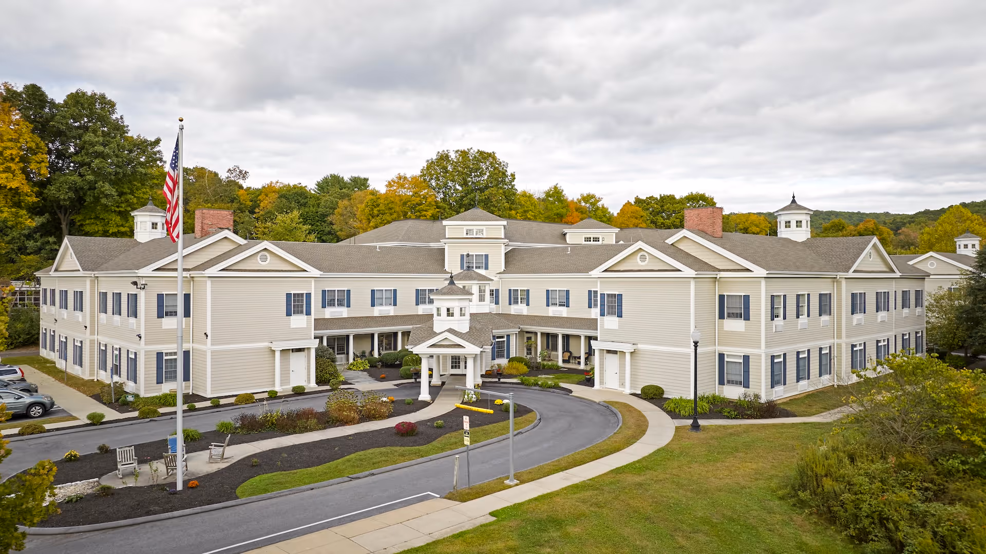 Exterior view of a large, two-story senior living facility building with beige siding, multiple windows with blue shutters, and a covered entrance with white columns. There is a circular driveway in front, landscaped greenery, an American flag on a flagpole, and trees in the background under a cloudy sky.