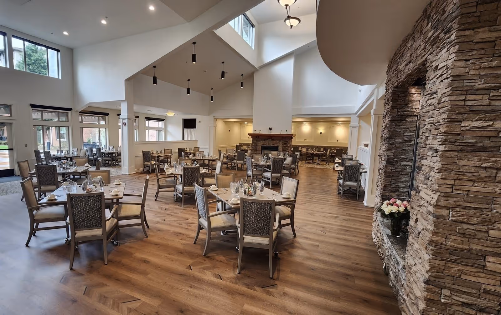 Spacious dining room with multiple tables and chairs arranged neatly on a wooden floor. The room features high ceilings with modern pendant lights, large windows allowing natural light, and a stone fireplace on the right side with a vase of flowers on the hearth.