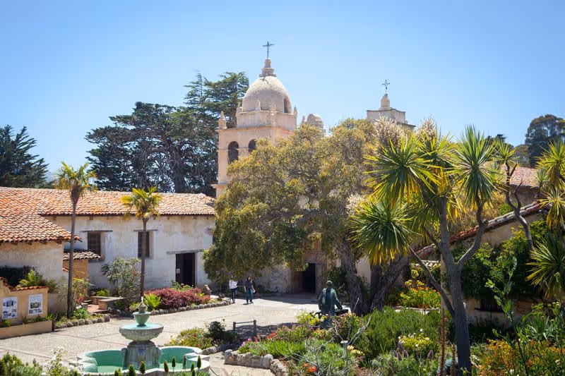 A sunny courtyard with a fountain in the foreground, surrounded by lush greenery and palm trees. In the background, there is a historic building with a bell tower topped with a cross. People are walking near the entrance of the building.