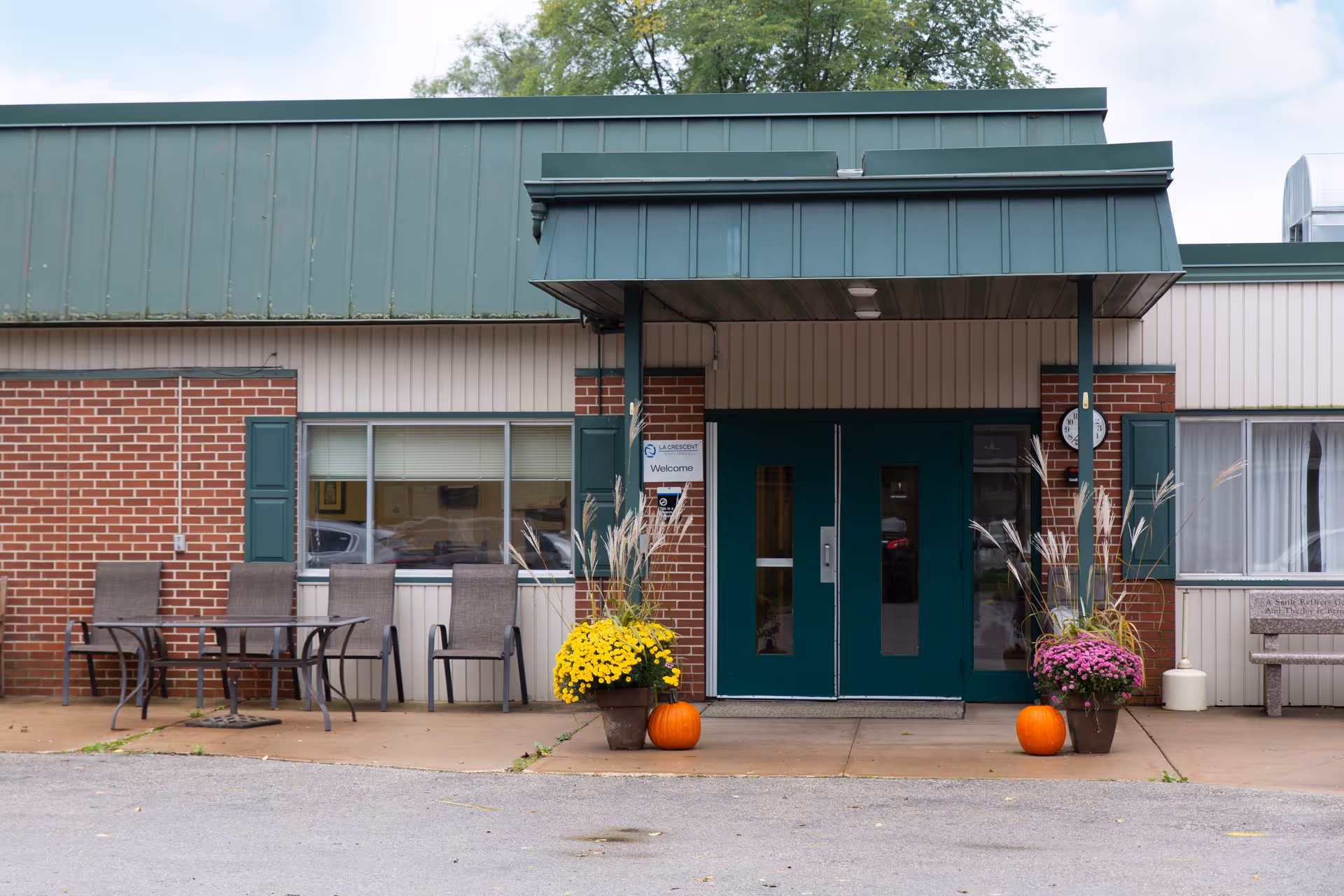 Front entrance of La Crescent Health Services building with green double doors, brick and beige siding, two large flower pots with yellow and purple flowers and pumpkins, outdoor seating with a table and chairs on the left, and a bench on the right.