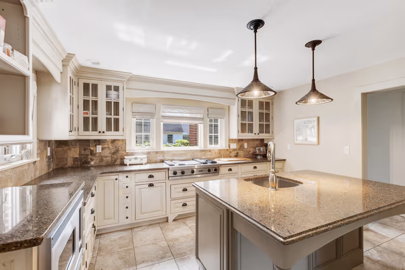 Bright kitchen with beige cabinets, granite countertops, a central island with a sink, two pendant lights hanging above the island, a stove under a window with three panes, and tiled flooring.
