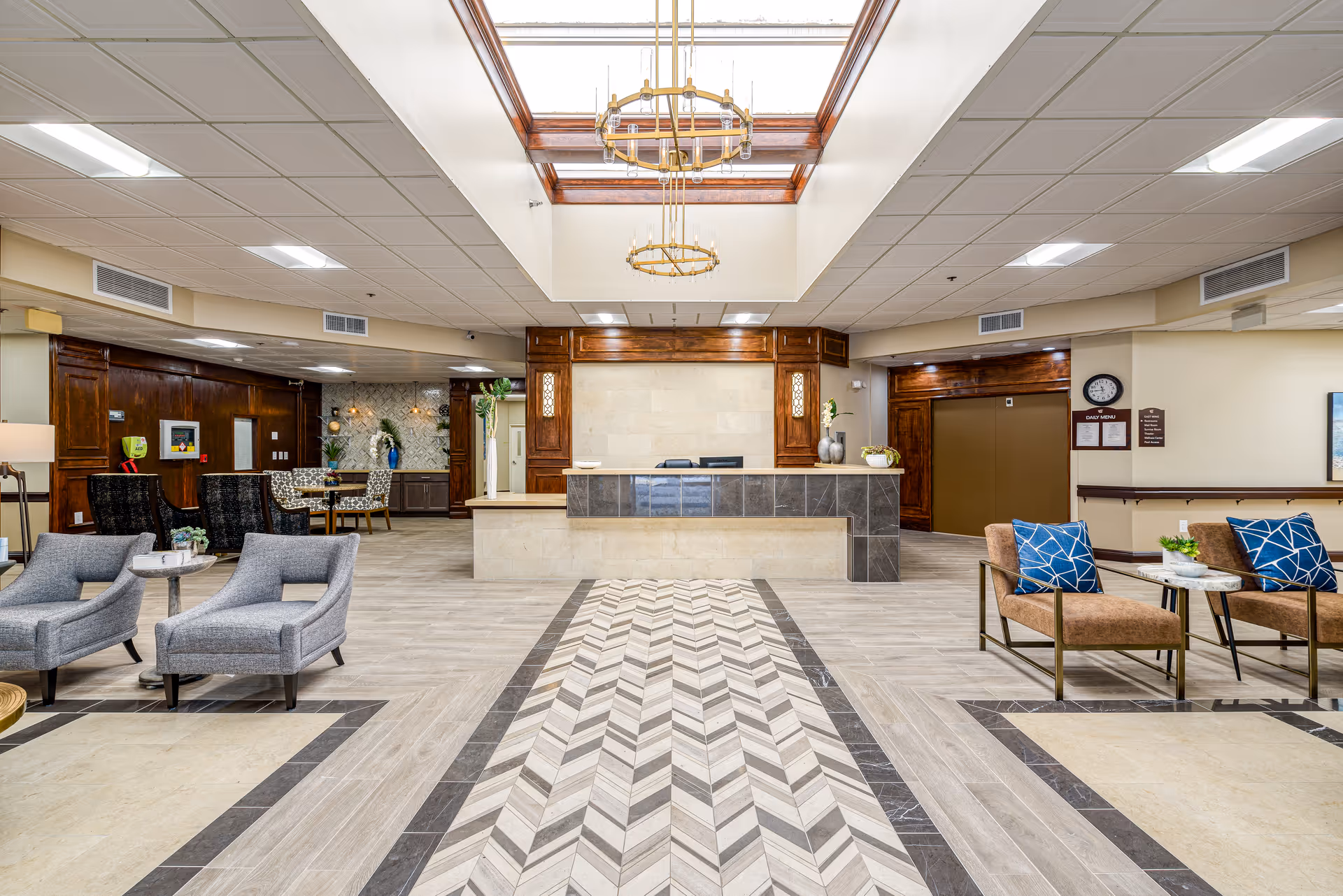 A spacious senior living facility lobby with a central reception desk made of beige and dark tiles. The ceiling features a large skylight with two hanging chandeliers. The floor has a patterned tile design leading to the desk. There are seating areas with modern chairs and small tables on either side of the lobby. The walls have wood paneling and light beige paint, with a clock and daily menu posted near an elevator on the right side.