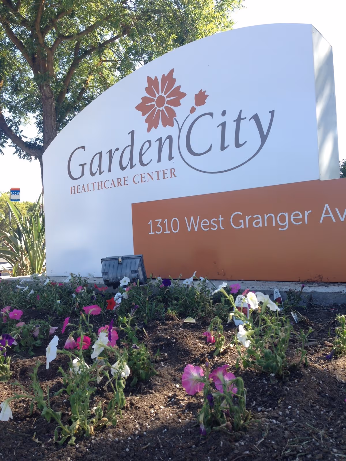 Outdoor sign for Garden City Healthcare Center with flowers planted in front and a tree in the background.