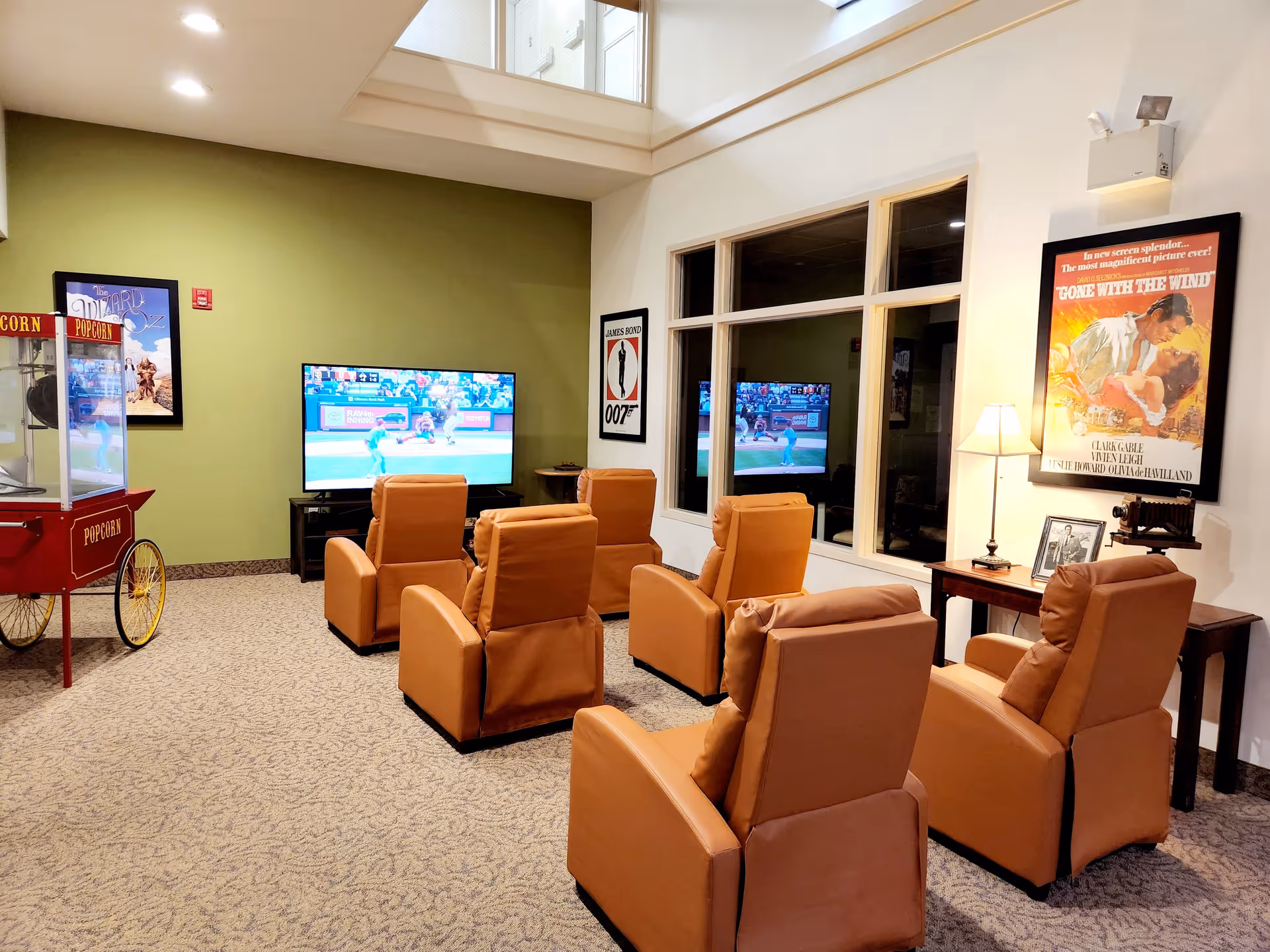 A cozy senior living facility media room with six tan leather recliners arranged in two rows facing a large flat-screen TV showing a baseball game. The room has a green accent wall with framed movie posters of The Wizard of Oz and James Bond 007. There is a vintage popcorn machine on the left and a wooden side table with a lamp, framed photo, and old camera on the right. A large window reflects the TV screen.