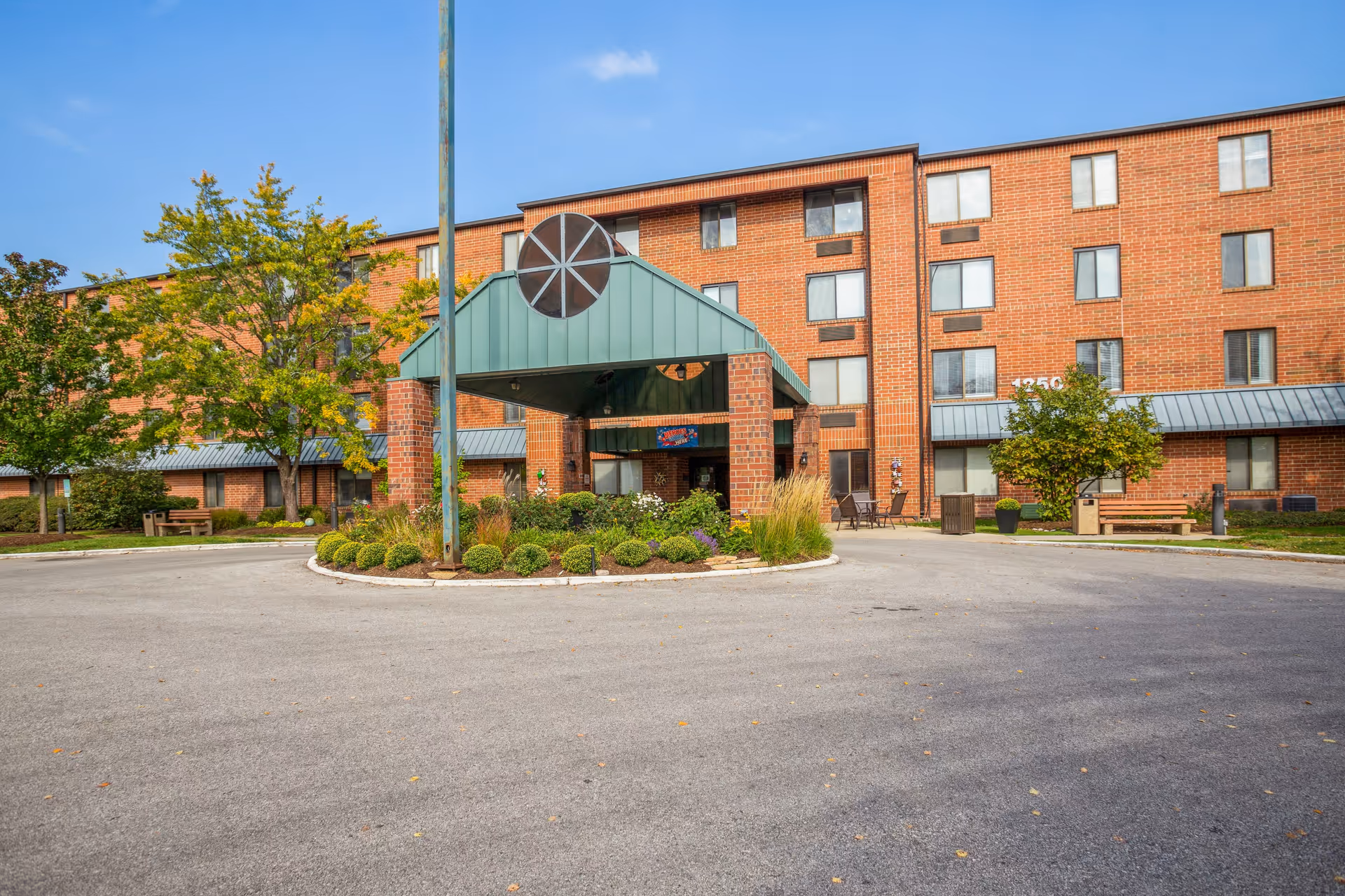 Front exterior view of a multi-story brick building with a covered entrance featuring a green metal roof and circular window design. There are landscaped bushes and trees around the entrance, benches, and outdoor seating areas visible. The sky is clear and blue.