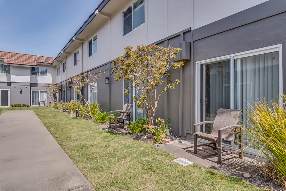 Outdoor view of Serra Highlands Senior Living showing a two-story building with sliding glass doors leading to small patios with chairs. There are small trees and plants along the building, and a concrete walkway runs alongside a grassy area.