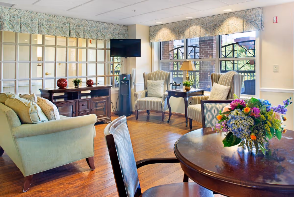 A bright and cozy living room area with a green sofa, two striped armchairs, a wooden side table with a lamp and decorative items, a wooden cabinet with plants and red decorative balls, a wall-mounted TV, a water dispenser, and large windows with patterned valances letting in natural light. A round wooden table with a colorful flower arrangement is in the foreground.