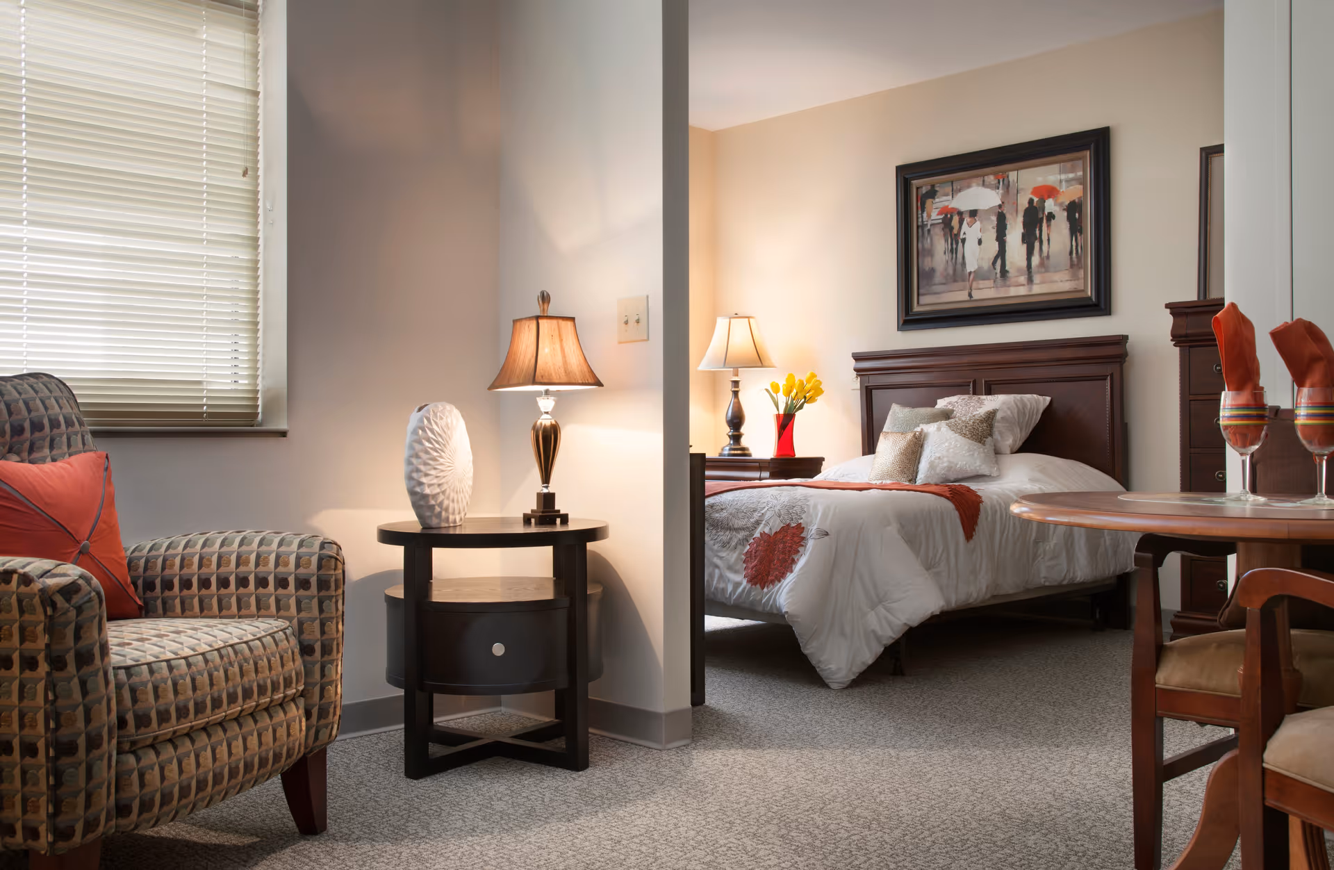 A cozy senior living facility room featuring a patterned armchair with a red pillow, a round side table with a decorative white vase and a lamp, and a bedroom area with a bed adorned with white and orange bedding, a nightstand with a lamp and yellow flowers, a framed painting of people with umbrellas on the wall, and a wooden dining table with two chairs and glasses with orange napkins.