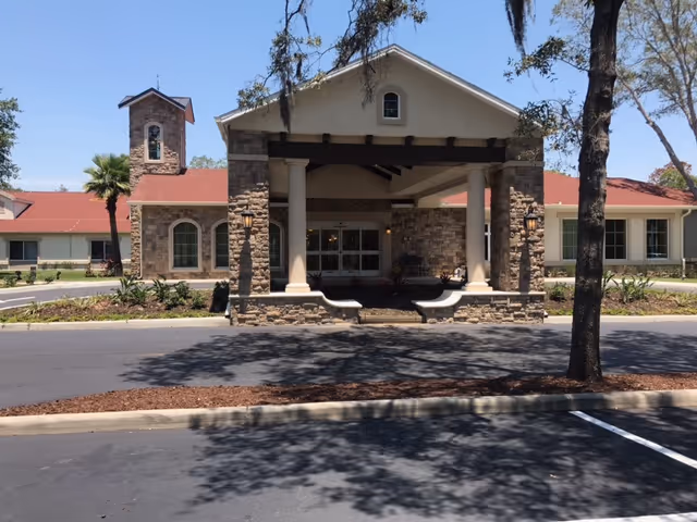 Front exterior view of a senior living facility building with stone and beige walls, a covered entrance supported by columns, a red roof, and a small tower-like structure on the left side. There are trees and landscaping around the building and a parking lot in the foreground.