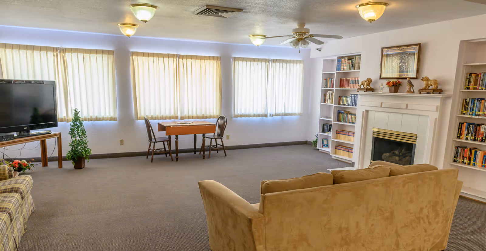 A spacious living room with beige carpeting, a tan sofa, a plaid armchair, a wooden table with two chairs near windows with closed curtains, a flat-screen TV on a wooden stand, a fireplace with decorative items on the mantel, and built-in bookshelves filled with books.