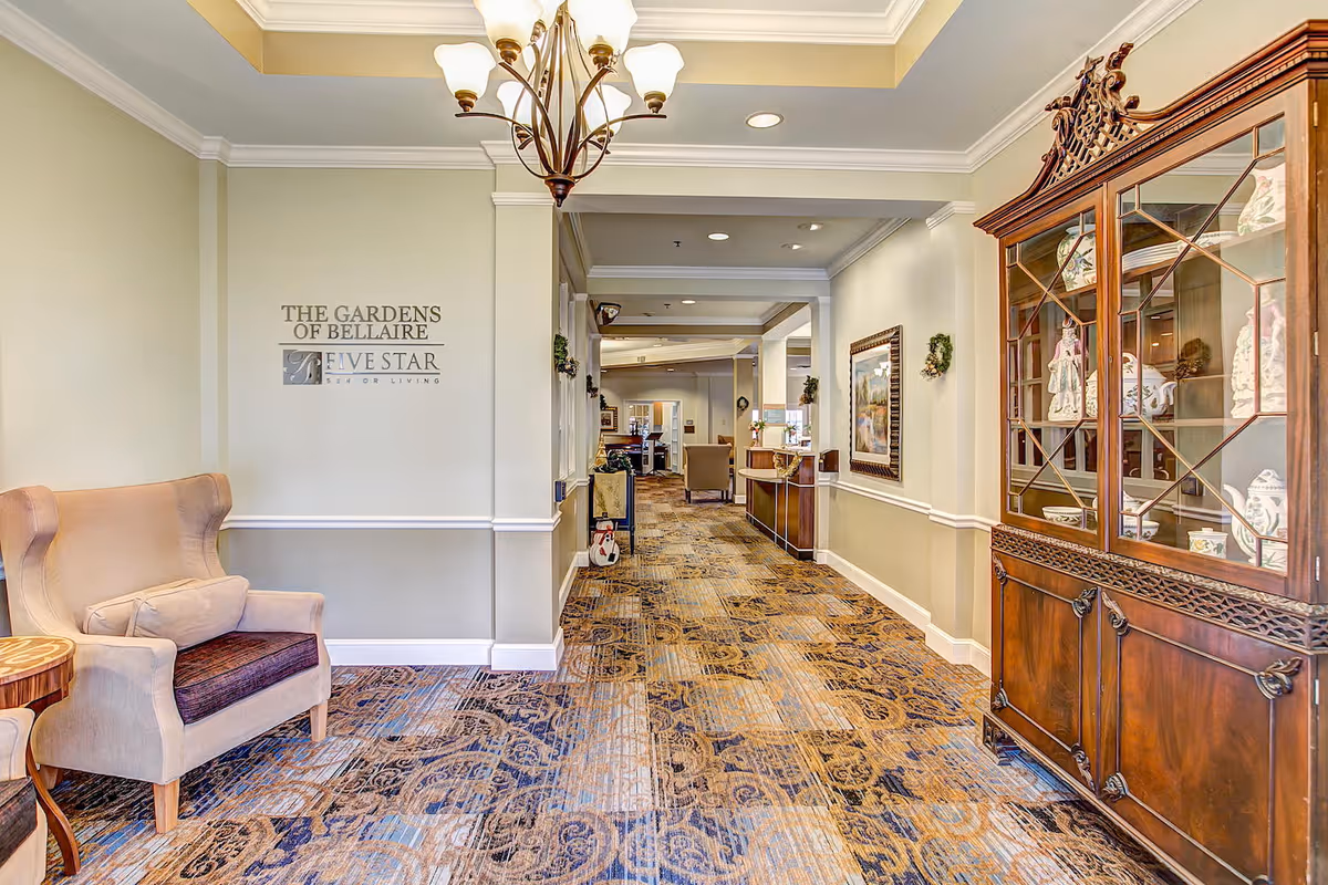Interior hallway of The Gardens of Bellaire senior living facility featuring a patterned carpet, beige walls with white trim, a chandelier, a beige armchair with a cushion, a wooden side table, and a wooden display cabinet with decorative items. The hallway leads to a reception area with seating and artwork on the walls.
