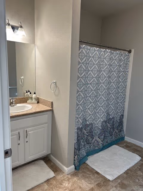 Bathroom with a white vanity cabinet, beige countertop, sink, and a mirror above. There is a wall-mounted light fixture with two bulbs above the mirror. A patterned shower curtain with blue and gray designs hangs on a curved shower rod. Two white bath mats are placed on the tiled floor, one in front of the vanity and one in front of the shower.