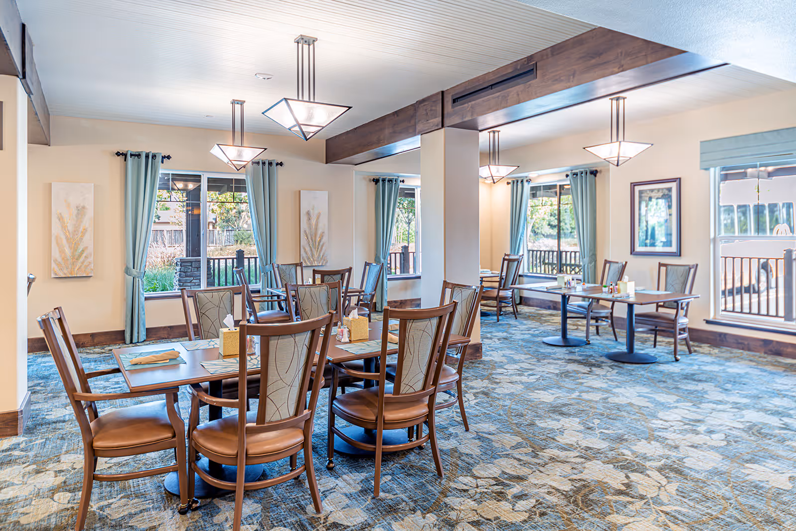 Bright, spacious dining room with multiple wooden tables and chairs, pendant lights, and large windows with blue curtains.