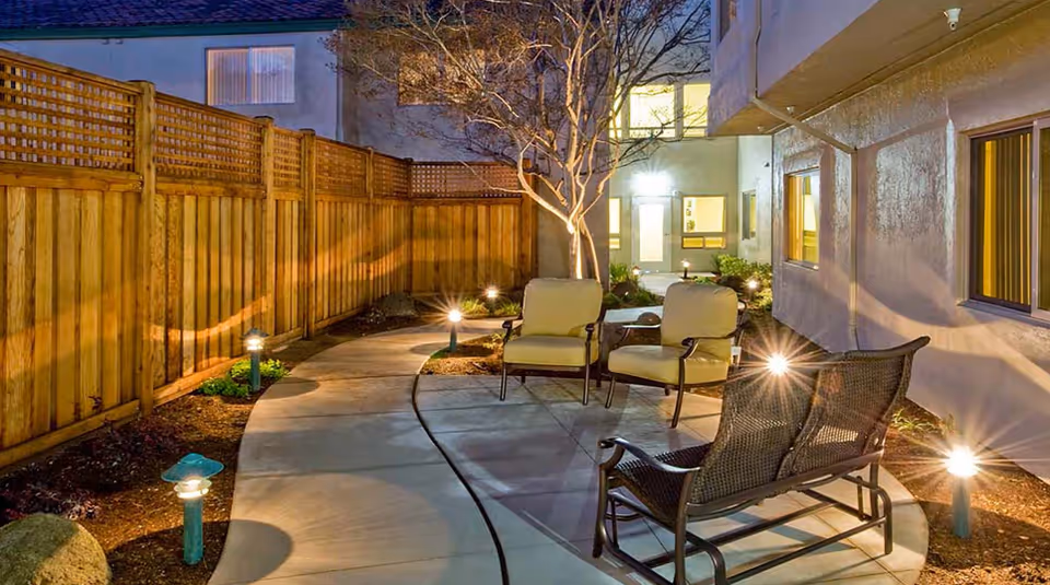 Outdoor patio area at night with cushioned chairs and a small table on a curved concrete pathway, surrounded by a wooden fence and illuminated by small ground lights. The building wall with windows is visible on the right side.