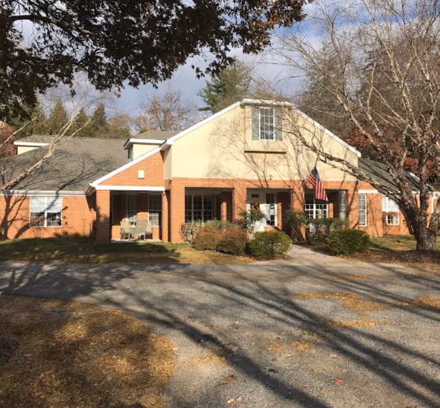 Front exterior view of a single-story senior living facility building with a brick and beige facade, an American flag near the entrance, surrounded by leafless trees and bushes under a clear sky.