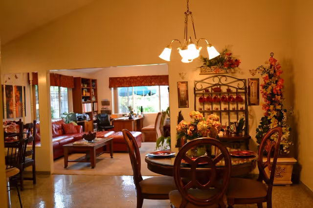 A warm and inviting interior space featuring a dining area with a round wooden table set with plates and surrounded by four chairs. The dining area is decorated with floral arrangements and a decorative wall rack holding red cups. In the background, there is a living room with a red leather sofa, a wooden coffee table, a desk with a chair, and large windows letting in natural light.