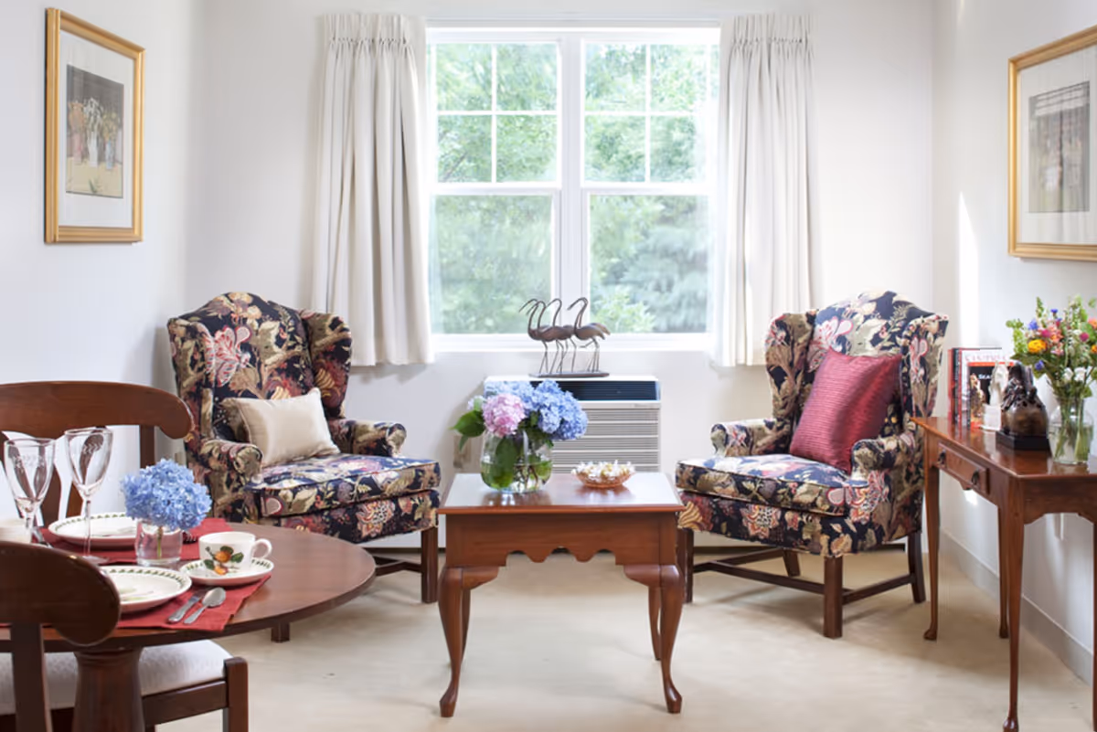 Sunlit sitting area with two floral wingback chairs facing a wooden coffee table and a small dining table set by a large window.