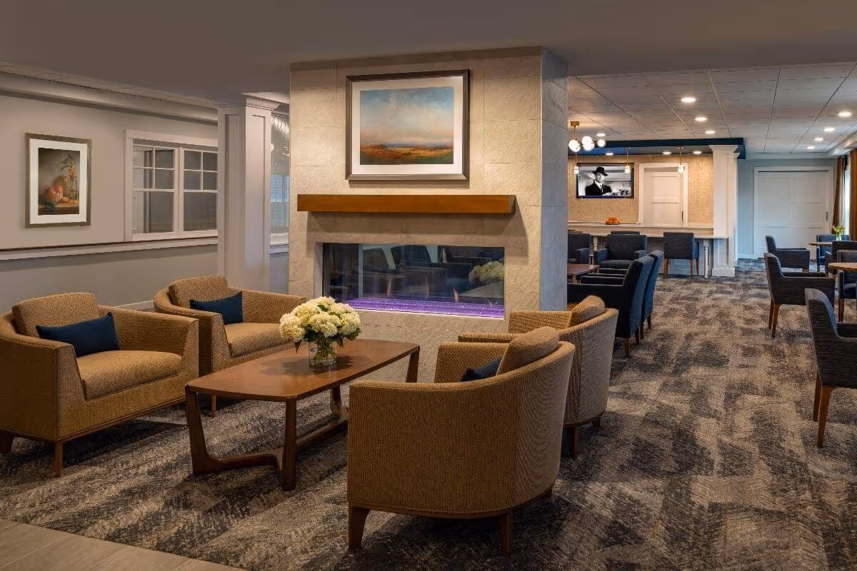 A cozy living room area in a senior living facility with four beige armchairs arranged around a wooden coffee table holding a vase of white flowers. A modern fireplace with a wooden mantle and a landscape painting above it is centered in the room. In the background, there are additional seating areas with dark chairs and tables, a television mounted on the wall, and soft overhead lighting.