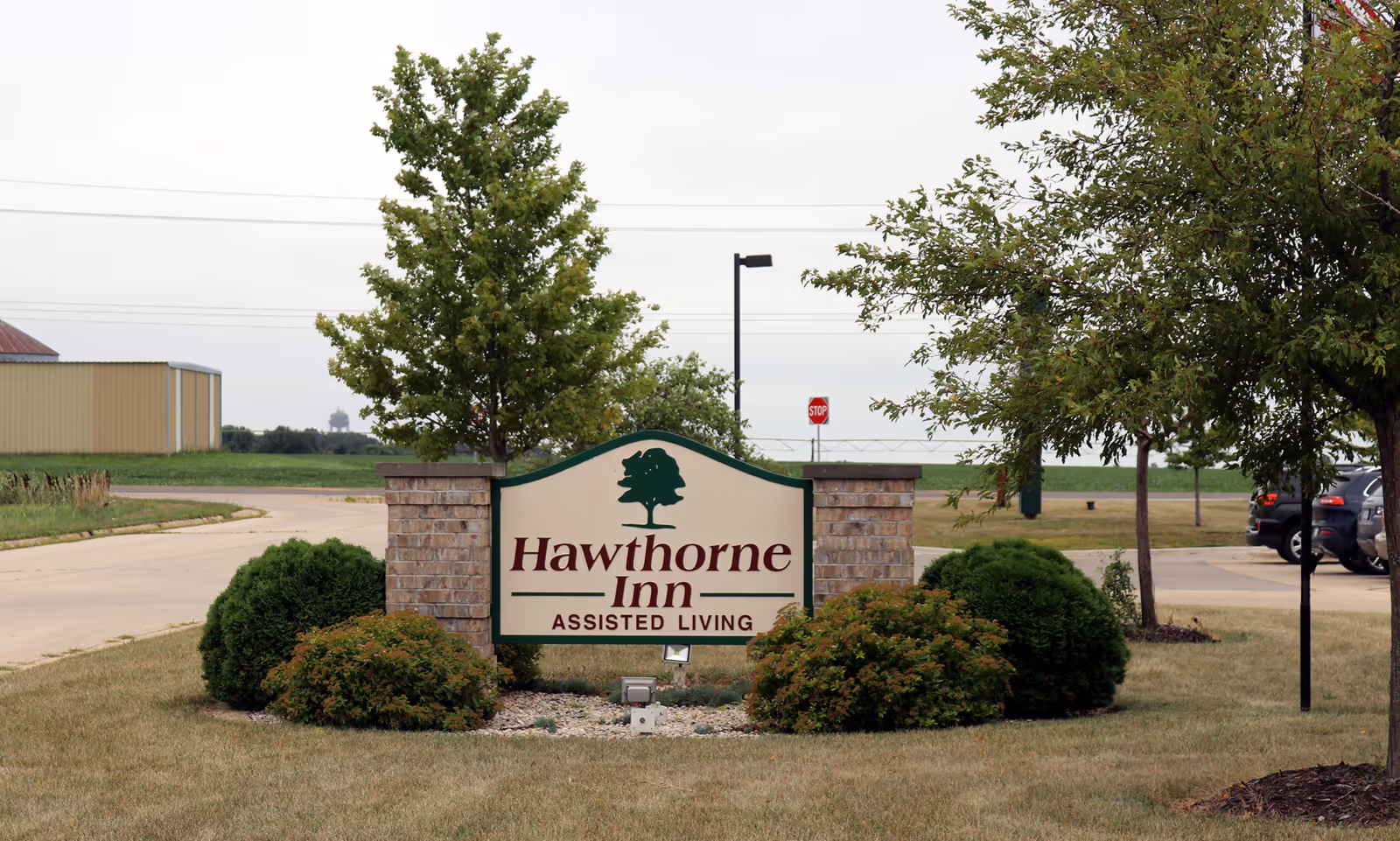 Outdoor view of a landscaped area with a sign that reads 'Hawthorne Inn Assisted Living' surrounded by bushes and trees, with a road and parked cars in the background.