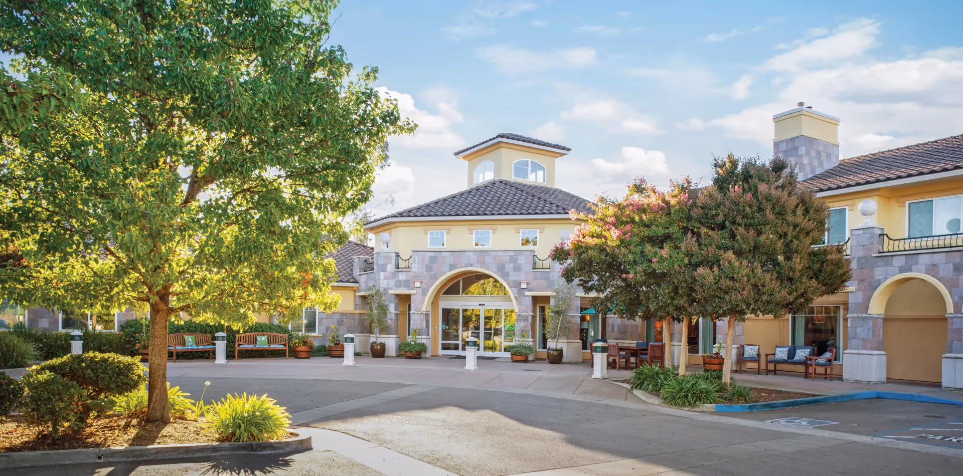 Exterior view of Cogir of Sonoma Plaza senior living facility with a circular driveway, trees, benches, and a building entrance featuring stone and stucco facade under a partly cloudy sky.