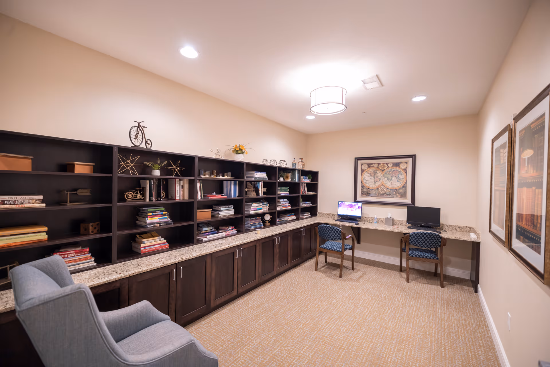 A quiet study or library room with built-in dark wood shelves filled with books and decorative items. There is a long granite countertop with two computer workstations, each with a chair. A comfortable armchair is positioned near the shelves. The walls are beige with framed artwork, and the room is softly lit by ceiling lights.