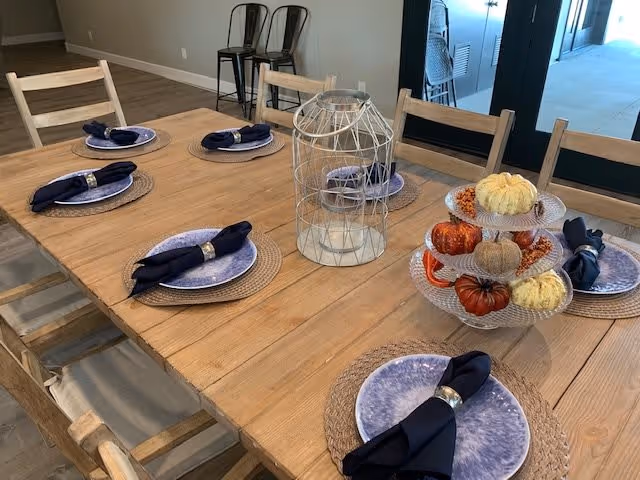 A wooden dining table set with six place settings, each with a blue plate, a woven placemat, and a navy blue cloth napkin with a silver napkin ring. The table also features a decorative white wire lantern and a three-tiered glass stand holding small decorative pumpkins in orange, white, and beige. In the background, there are two black metal chairs against a beige wall and a glass door leading to another room.