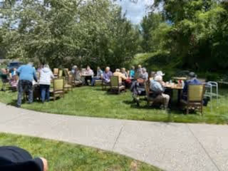 A group of elderly people sitting on chairs arranged in a circle on a grassy lawn, surrounded by trees and greenery, enjoying an outdoor gathering on a sunny day.