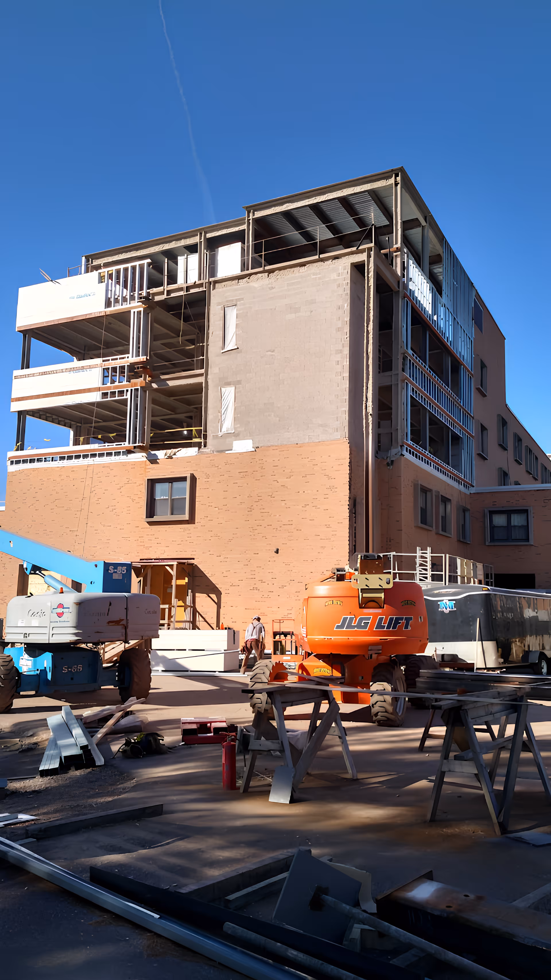 A multi-story building under construction with exposed steel framing and partially completed walls. Construction equipment, including a blue lift and an orange JLG lift, are parked in front of the building. Construction materials and tools are scattered around the site under a clear blue sky.