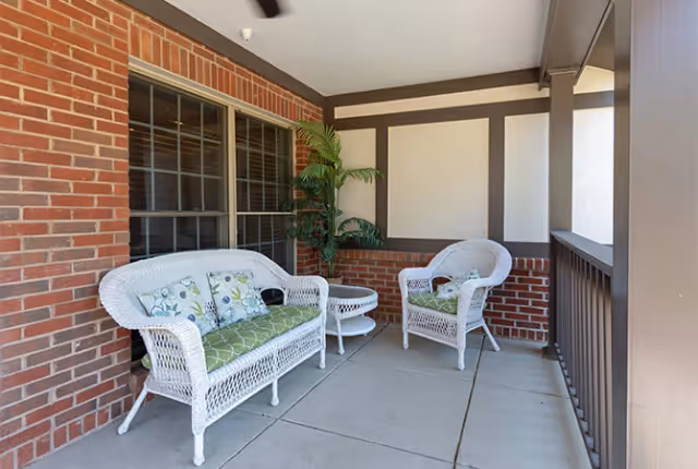 A covered outdoor patio area with white wicker furniture including a loveseat, a chair, and a small round table. The loveseat and chair have green cushions with floral patterned pillows. The patio has brick walls and a concrete floor, with a potted plant in the corner.