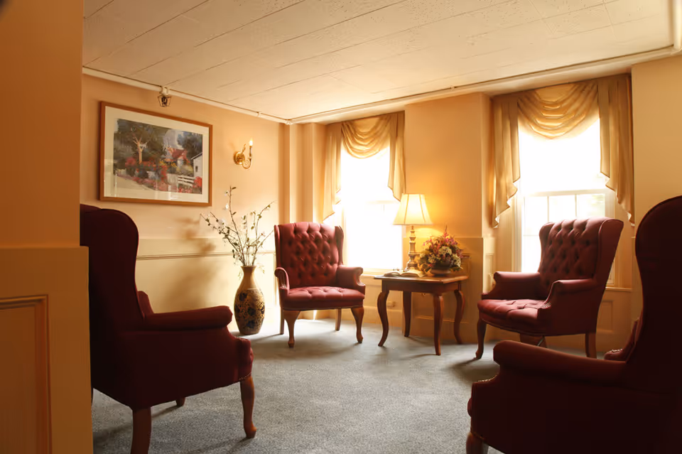 A cozy sitting area with four burgundy upholstered armchairs arranged around a wooden side table with a floral arrangement and a lamp. The room has beige walls, two large windows with golden drapes, a framed painting on the wall, and a decorative vase with branches.