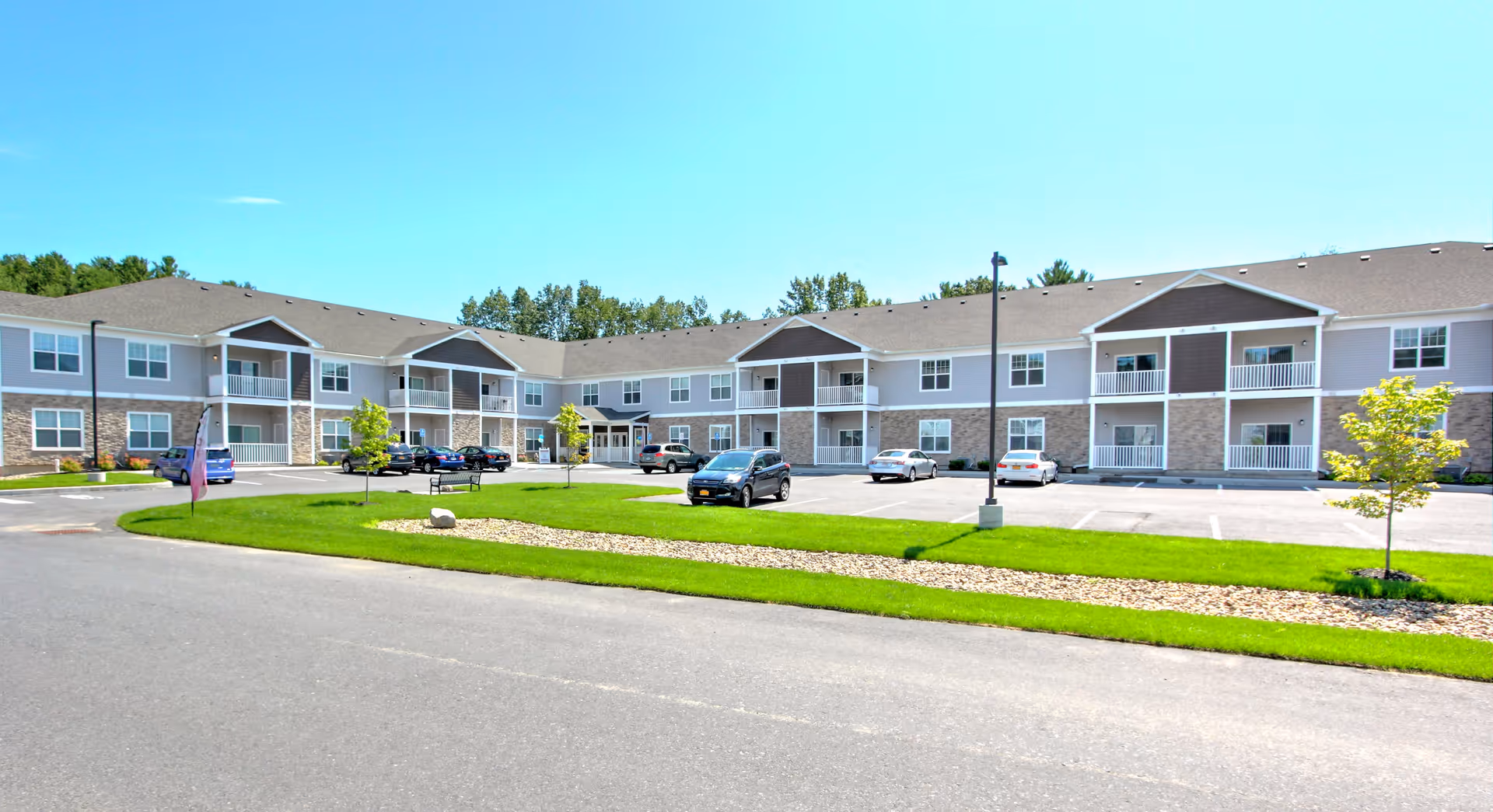 Exterior view of Carlton Hollow Senior Apartments, a two-story building with balconies, multiple windows, and a parking lot with several cars parked. The building is surrounded by green grass, small trees, and a clear blue sky.
