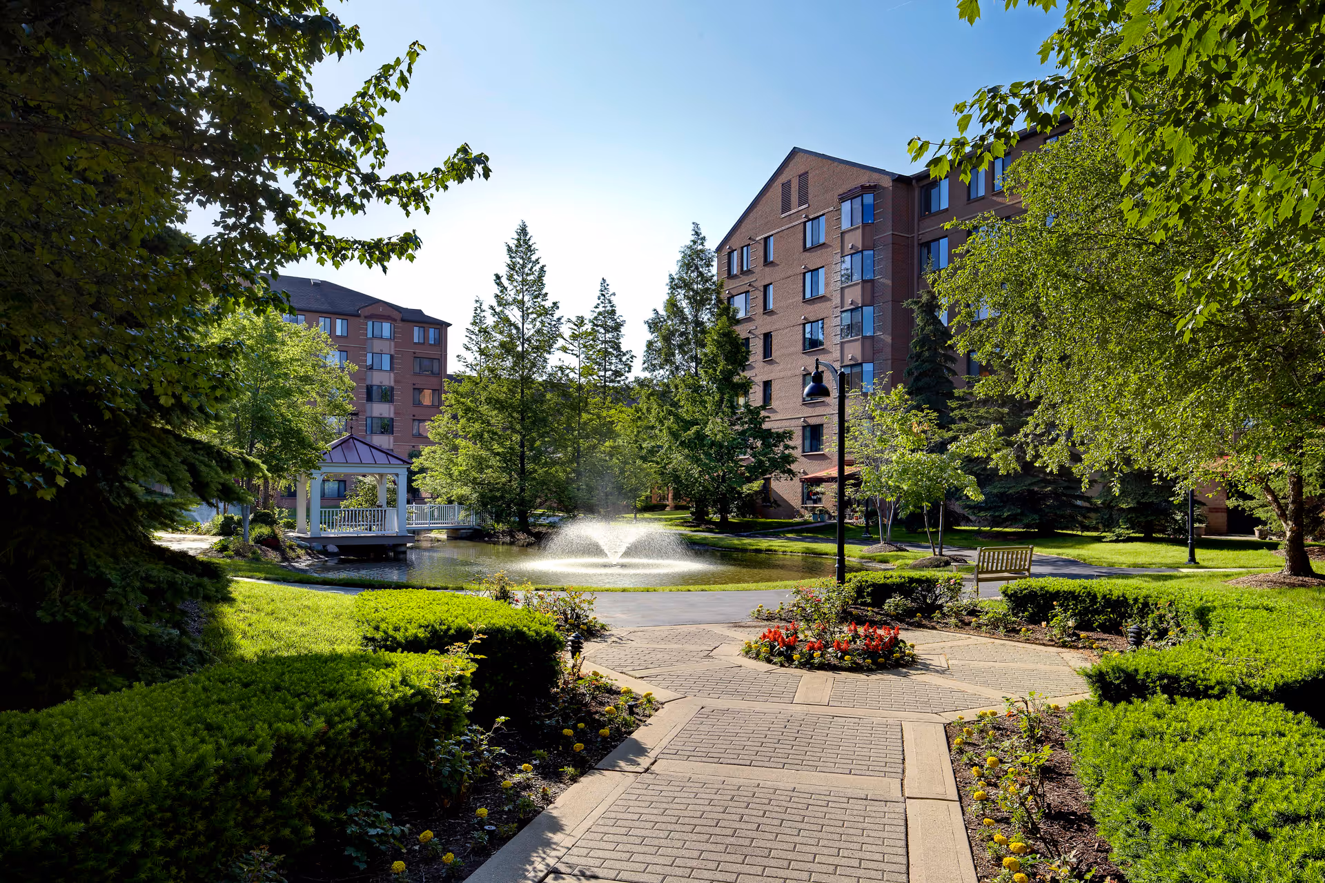 Landscaped courtyard featuring a pond with a fountain, a gazebo, paved walkways, benches, and multi-story brick buildings in the background.