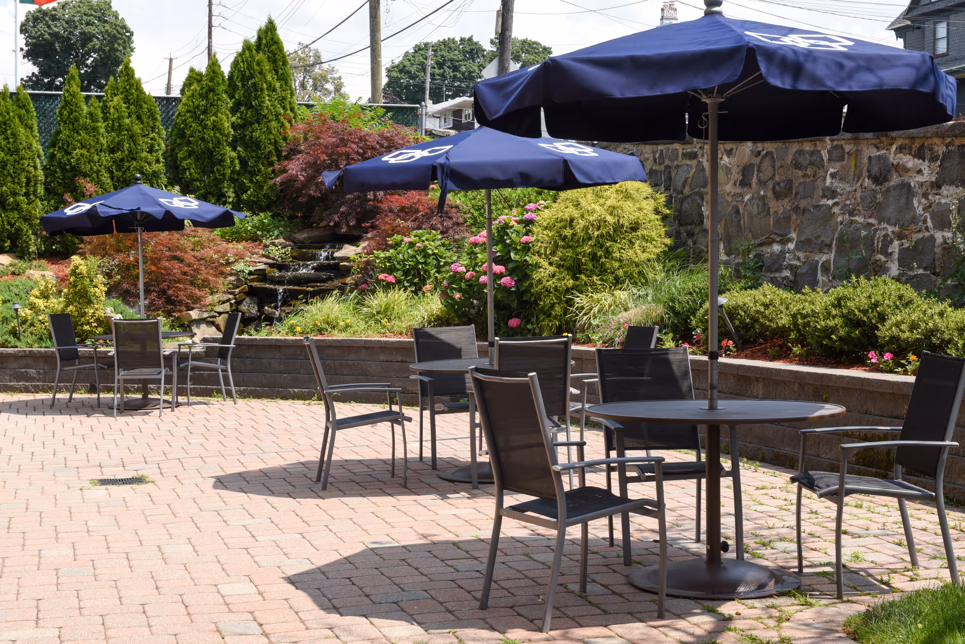 Outdoor patio area with several round tables and chairs, each table shaded by a navy blue umbrella. The patio is paved with bricks and surrounded by a stone wall and lush greenery including bushes, trees, and flowering plants.