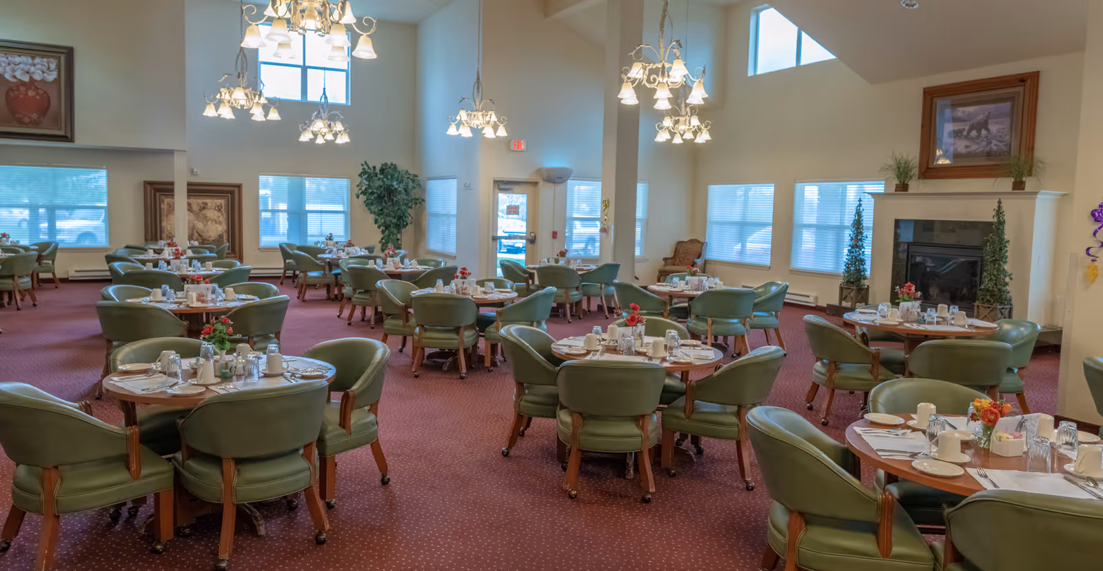 A spacious dining room with multiple round tables set with plates, cups, and silverware. Each table is surrounded by green cushioned chairs on wheels. The room has high ceilings with several chandeliers, large windows letting in natural light, framed artwork on the walls, and a fireplace with decorative plants on either side.
