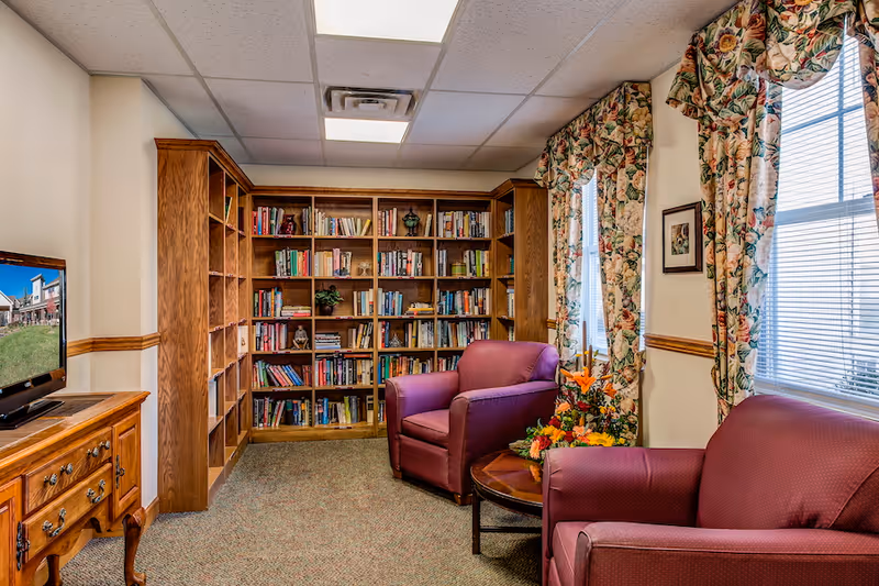 A cozy reading room with wooden bookshelves filled with books, two maroon armchairs, a wooden coffee table with a floral arrangement, floral curtains on the windows, and a wooden cabinet with a television on top.