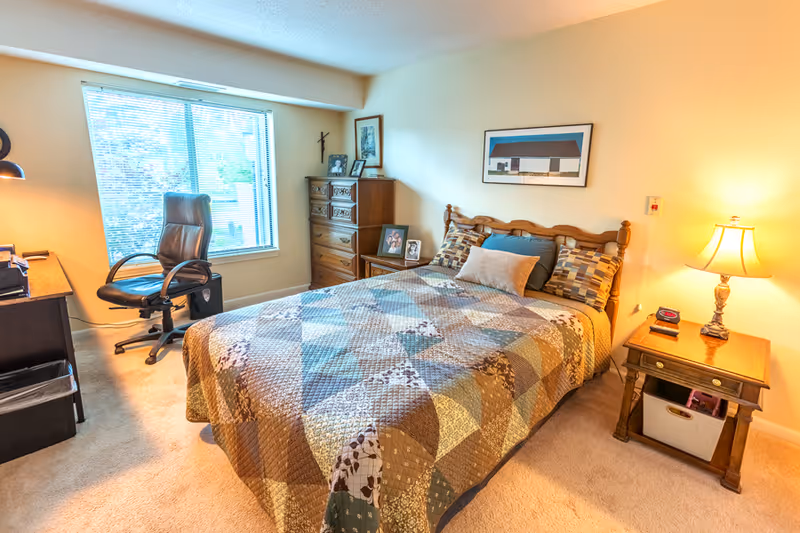 A cozy bedroom with a large window letting in natural light, a wooden bed with a patchwork quilt, a nightstand with a lamp and clock, a wooden dresser with framed photos, and a black office chair next to a desk.