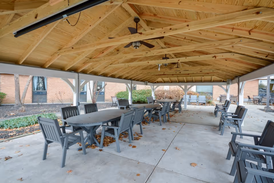 Covered outdoor seating area with multiple gray tables and chairs under a wooden roof structure. The area is paved with concrete and surrounded by some landscaping and brick building walls.