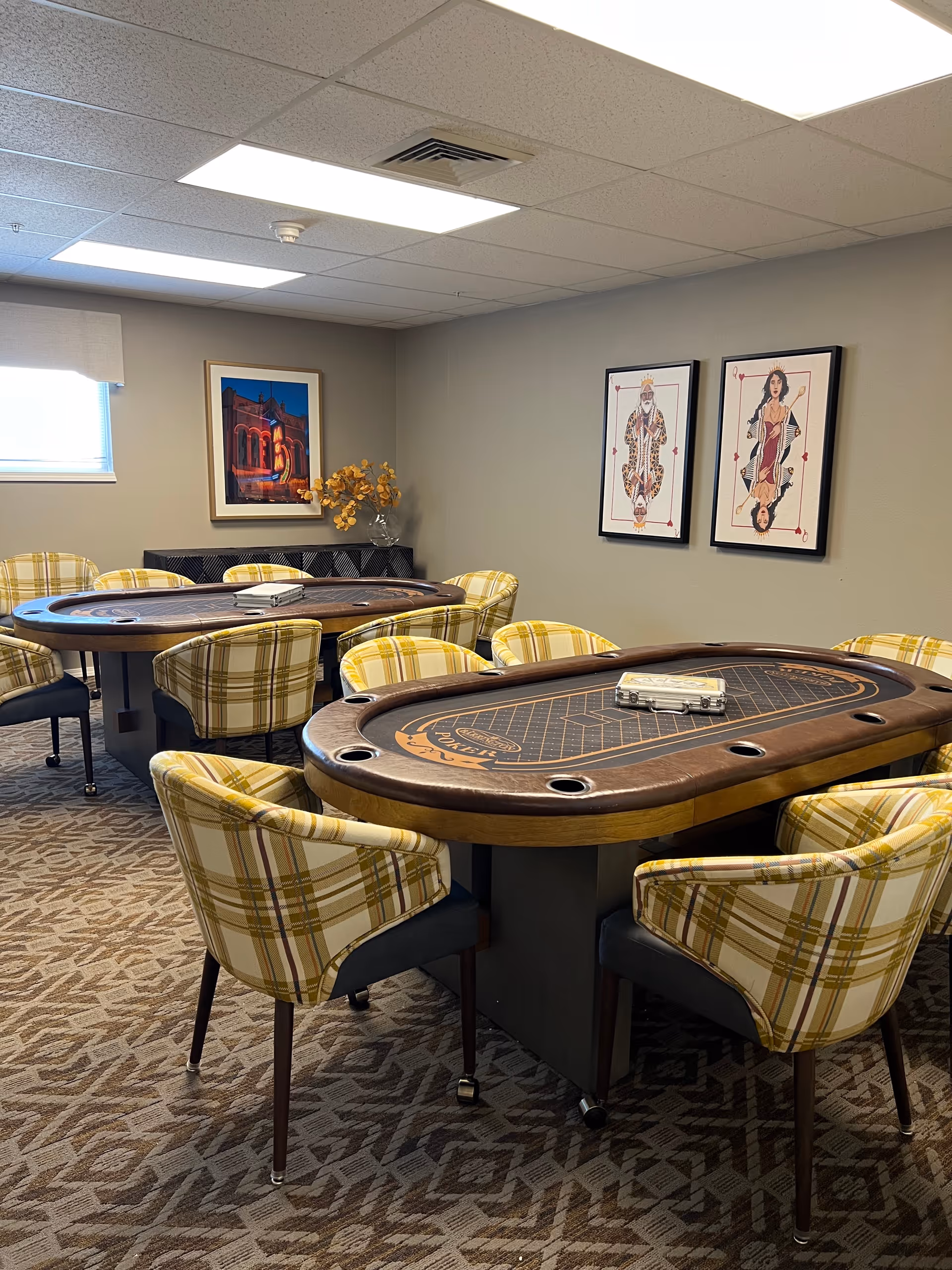 A room with two poker tables surrounded by yellow plaid upholstered chairs. The room has patterned carpet, a window with a blind, and framed artwork on the walls including two playing card themed pictures and one architectural photo. A vase with yellow flowers sits on a black cabinet against the wall.