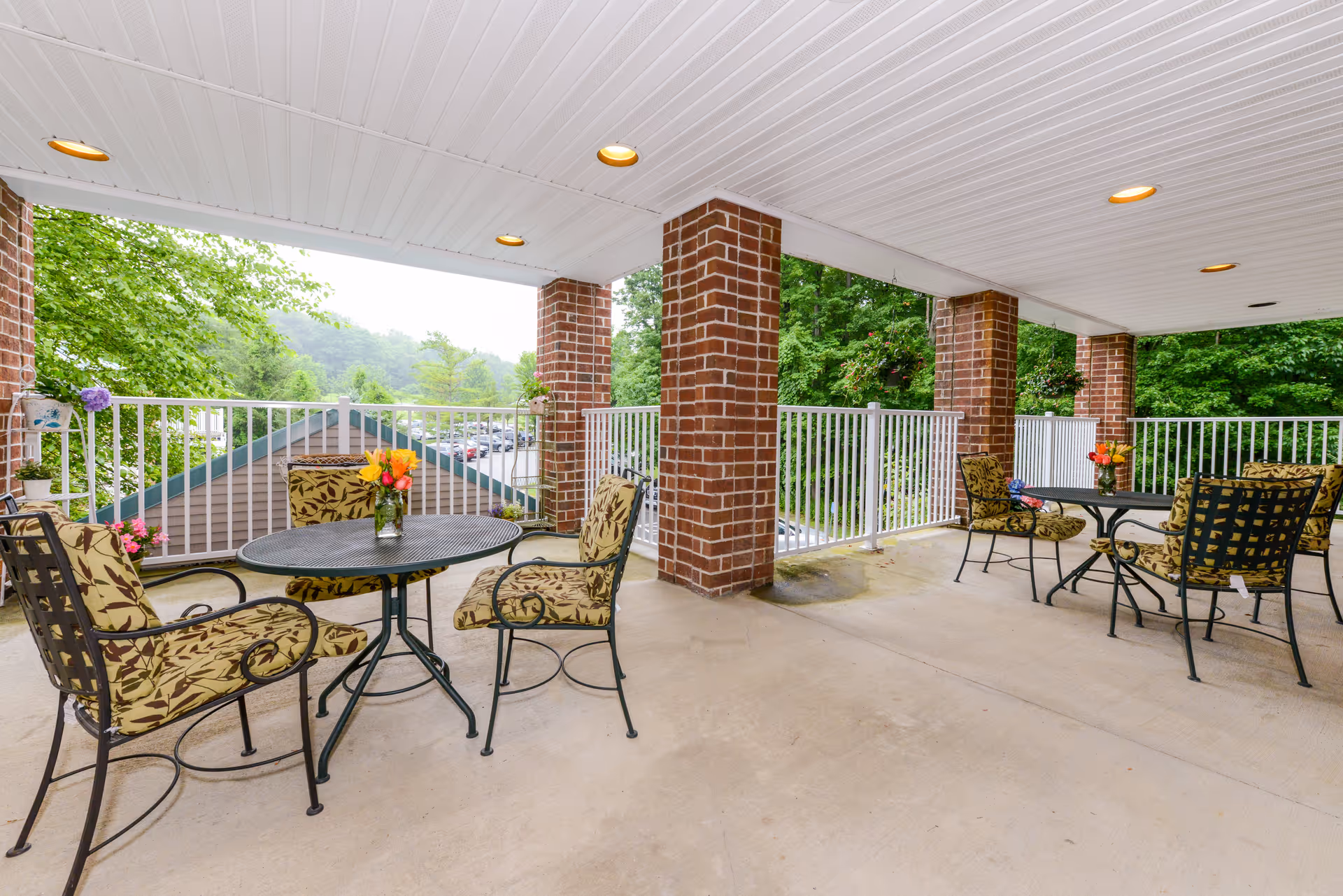 Covered outdoor patio area with metal tables and cushioned chairs featuring floral patterns. The patio has brick pillars, white railings, and a white ceiling with recessed lighting. There are flower vases on the tables and hanging plants along the railing. Trees and a parking lot are visible in the background.