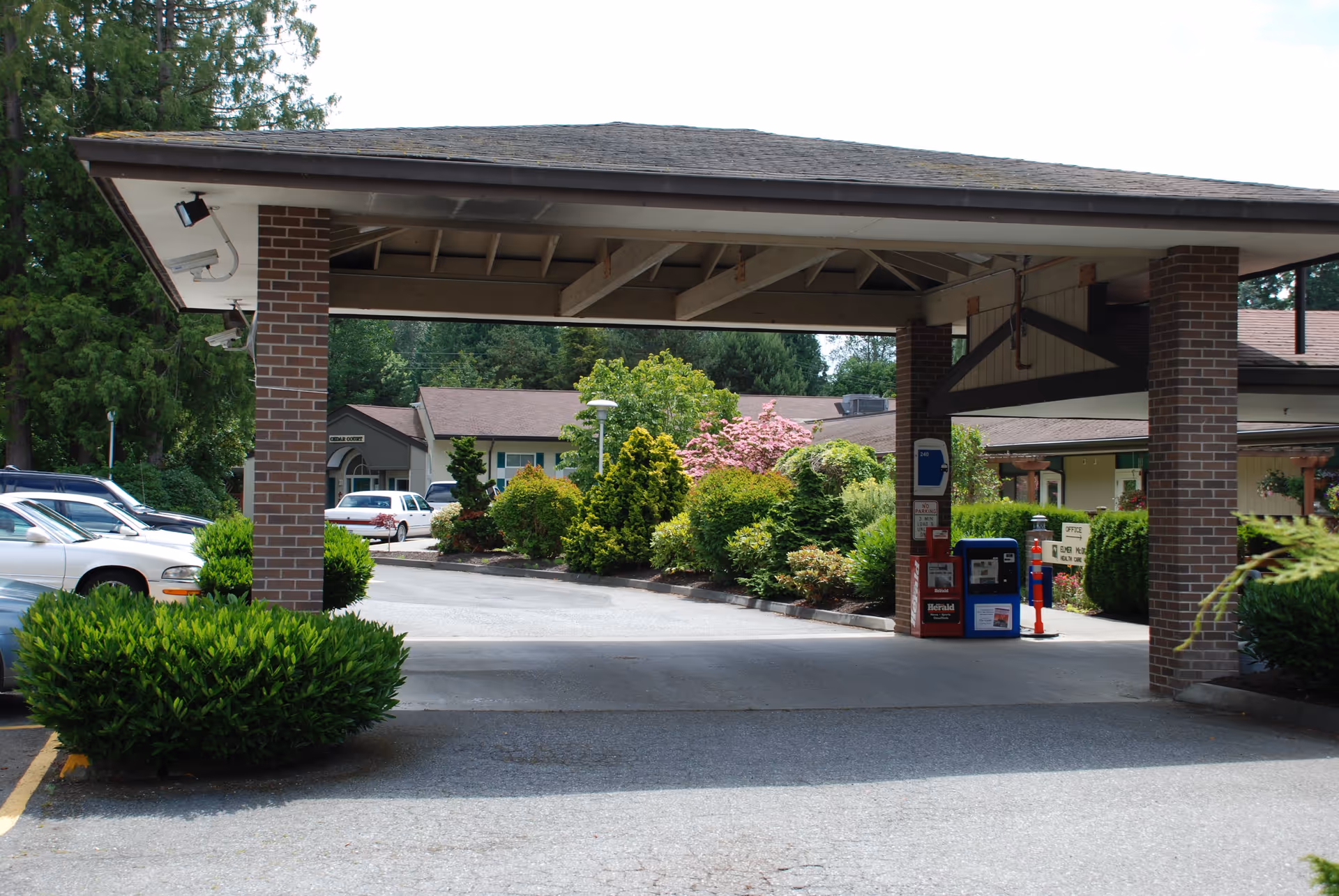 Covered driveway entrance with brick pillars at Warm Beach senior community, surrounded by green bushes and trees, with parked cars visible on the left and a building in the background.