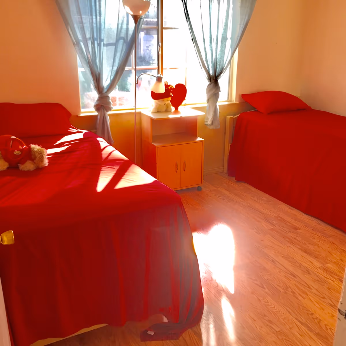 A bright bedroom with two single beds covered in red bedspreads. Between the beds is a small wooden nightstand with a white lamp, a stuffed animal, and a red heart decoration. Light blue curtains hang on the window behind the nightstand, and sunlight streams into the room, casting shadows on the wooden floor.