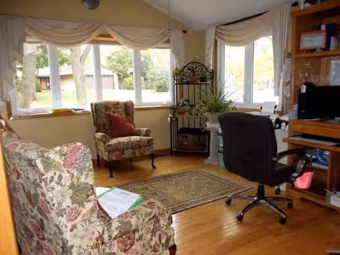Cozy senior living room with floral chairs, window drapes, desk, and potted plants.