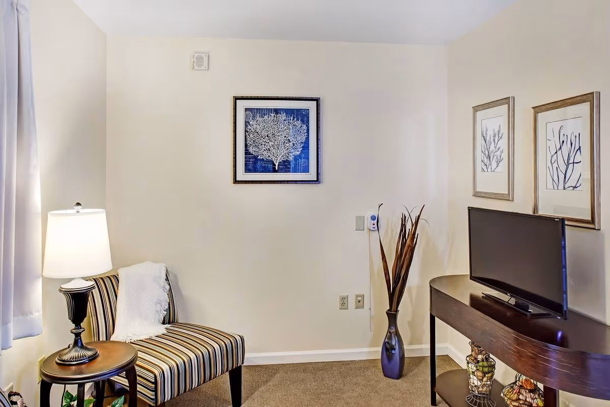 A small sitting area with a striped chair next to a round wooden side table holding a lamp. On the wall above the chair is a framed picture of a white coral on a blue background. To the right, there is a dark wooden console table with a flat-screen TV on top and decorative items underneath. Two framed pictures of coral are hung on the wall above the TV. A tall vase with dried branches is placed between the chair and the console table. The room has beige walls and carpeted floor.
