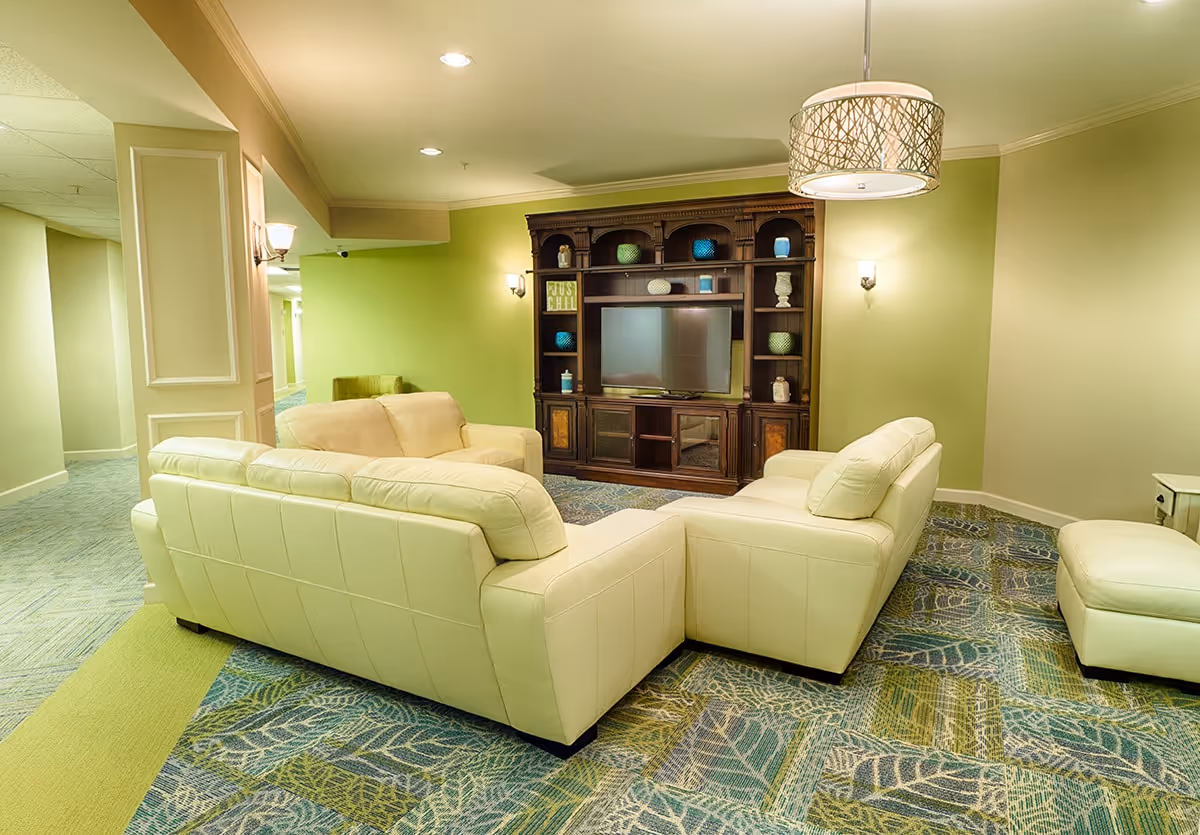 A cozy living room area with cream-colored leather sofas arranged around a large dark wooden entertainment center holding a flat-screen TV and decorative items. The room has light green walls, patterned carpet with leaf designs, and soft lighting from wall sconces and a hanging lamp.