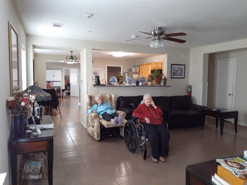 A living room area in an assisted living facility with two elderly women, one sitting in a floral armchair and the other in a wheelchair. The room has a ceiling fan, a black leather couch, a side table, and a kitchen area visible in the background. There are decorative items on the kitchen counter and a hallway leading to other rooms.